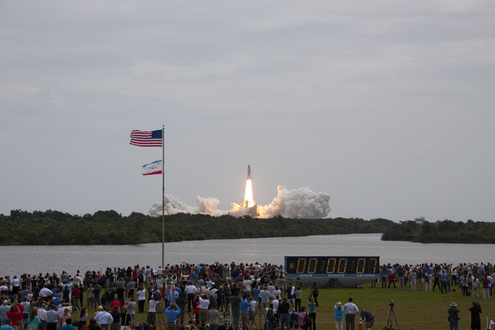 L'iconico orologio posto all'esterno della sala stampa del KSC è stato testimone di buona parte della storia dell'satronautica. In questa immagine il lancio dell'ultima missione del programma Space Shuttle, l'STS-135, decollata dal KSC l'8 Luglio 2011. (C) KSC/NASA